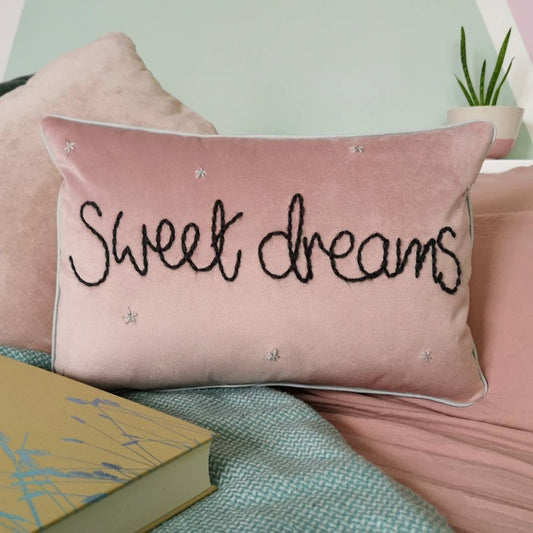 Pink pillow with 'Sweet dreams' text on a bed with a book and plant in the background