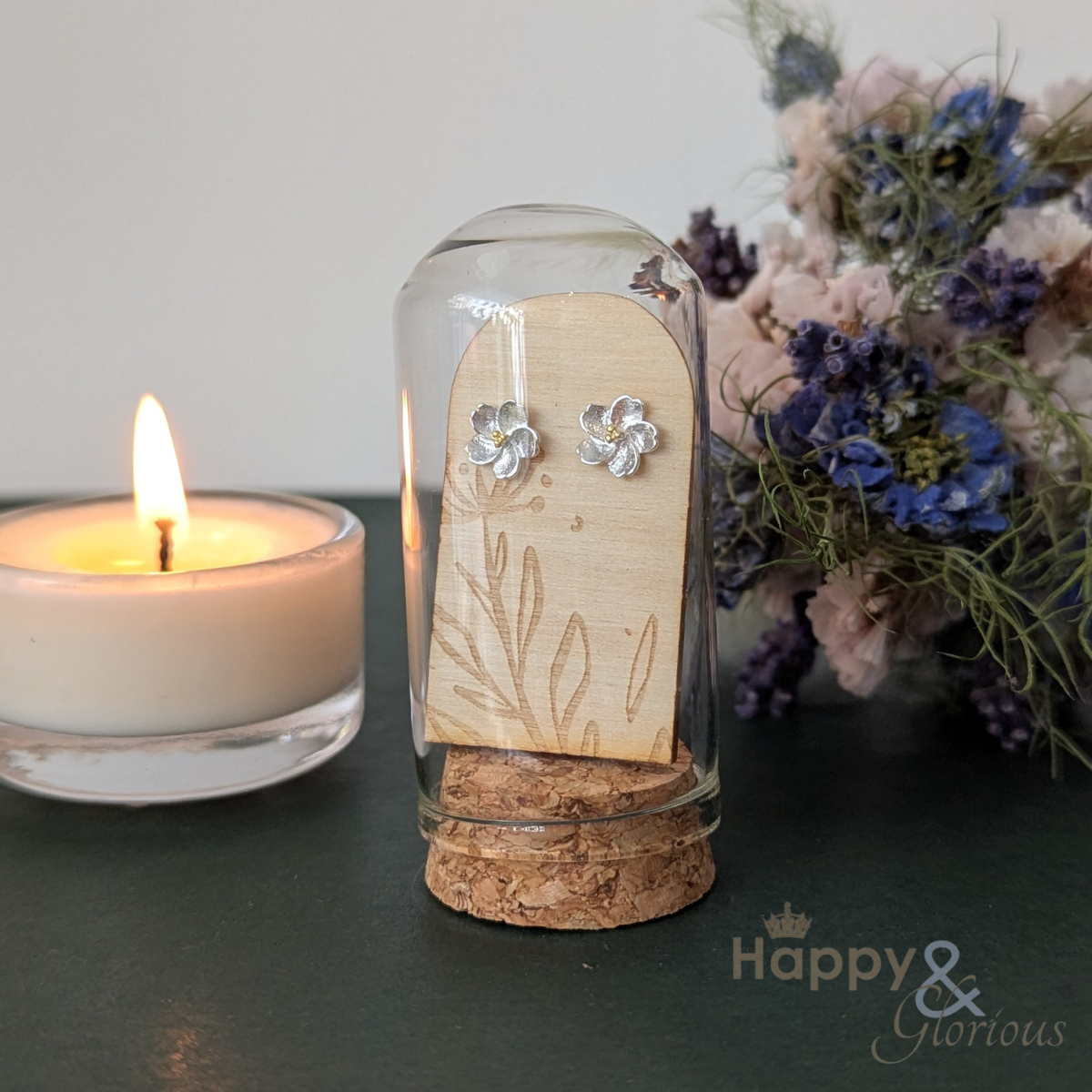 Glass container with wooden insert and earrings, lit candle and flowers in the background