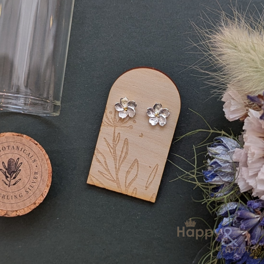 Flower-shaped silver earrings on a wooden display card with Botanicaus branding, surrounded by flowers on a dark surface.