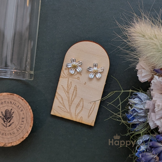 Flower-shaped earrings on a wooden display card with dried flowers and a glass container in the background.