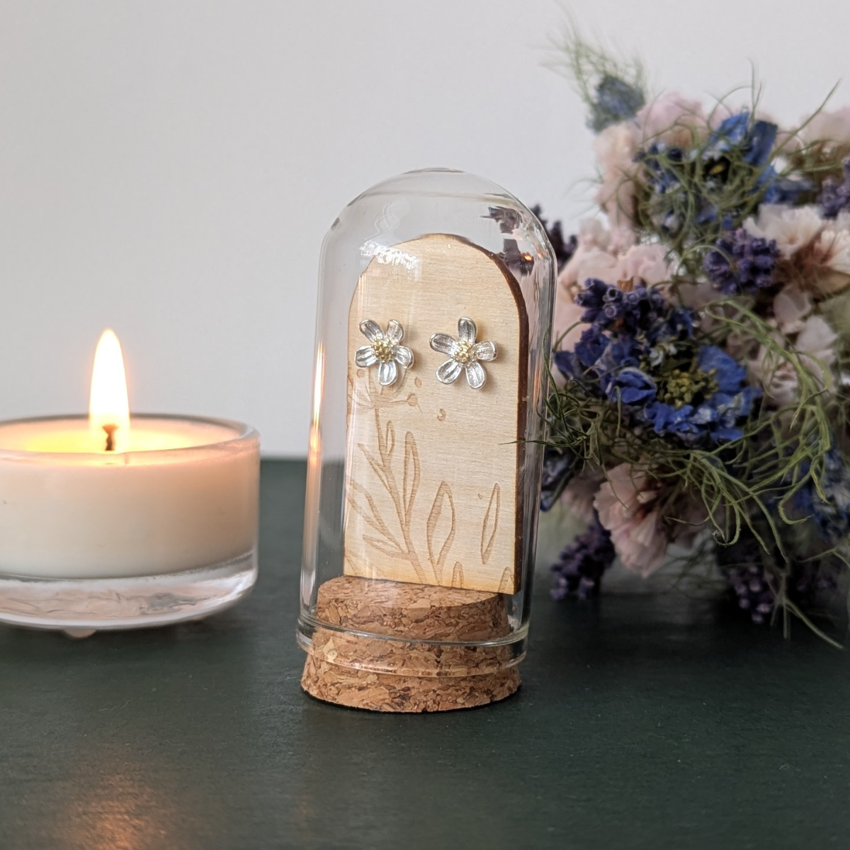 Pair of floral earrings in a glass dome with a lit candle and flowers in the background