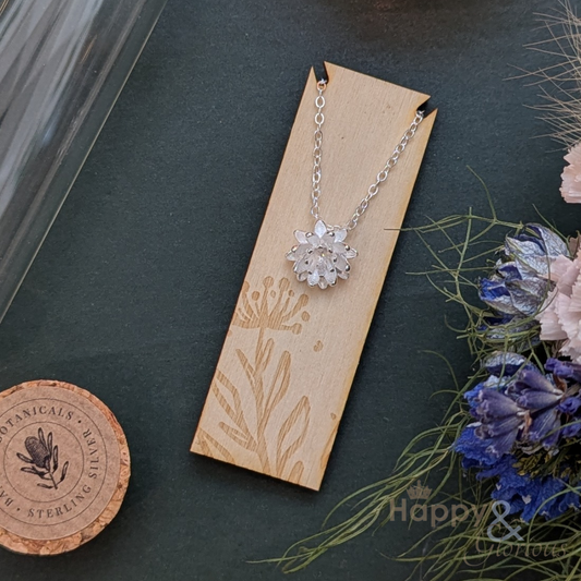 Silver necklace with a floral pendant on a wooden card, surrounded by dried flowers on a dark surface.
