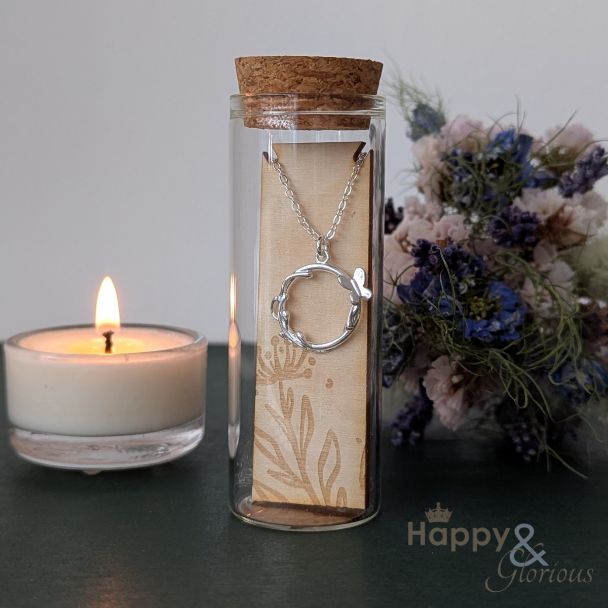 Silver necklace in a glass jar with a cork lid, next to a lit candle and flowers on a dark surface.