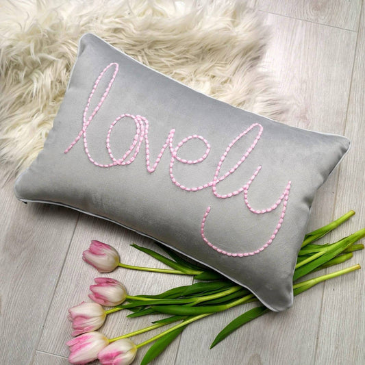 Gray pillow with 'lovely' embroidery surrounded by pink tulips on a wooden surface.