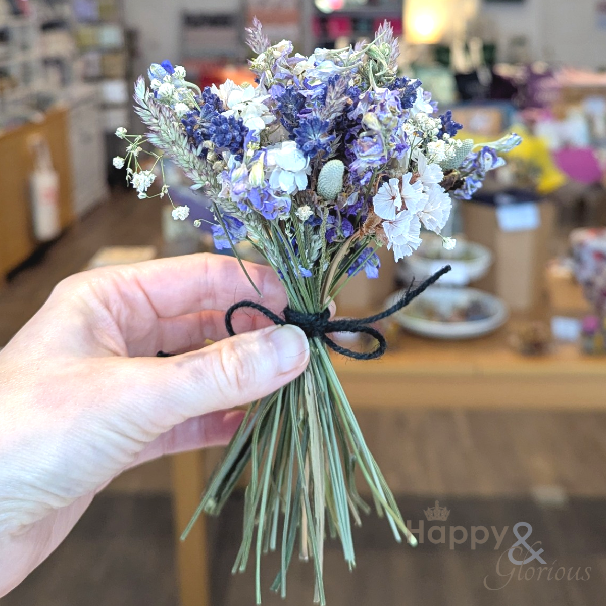 Hand holding a small bouquet of purple and white flowers with a blurred indoor background.