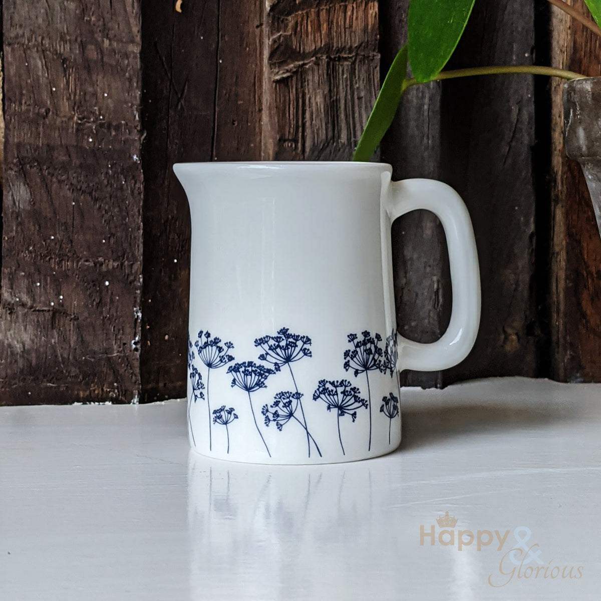 A fine bone china jug with a navy blue and white cow parsley pattern, designed by Kate Tompsett, placed on a white surface with a wooden wall in the background.