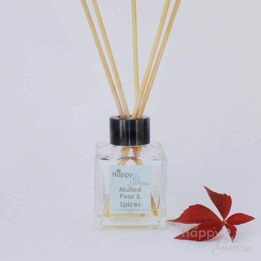 A clear glass diffuser bottle with a black cap, containing clear liquid and displaying labels with 'mulled pear & spices'. It has several light brown rattan reeds inserted, and a red leaf is placed to the side on a white background.