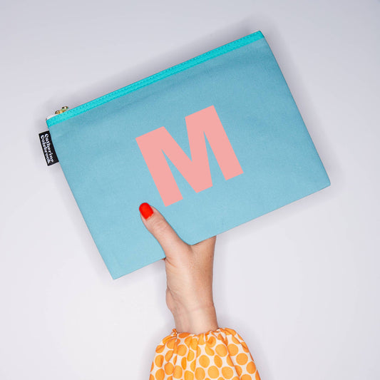 Light blue pouch with pink 'M' held by a hand with red nail polish on a light gray background