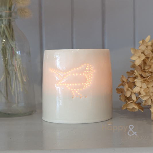 Ceramic candle holder with decorative light inside on a surface with flowers and a jar in the background.