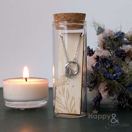 Necklace in a glass bottle with a cork lid, next to a lit candle and flowers on a gray surface.
