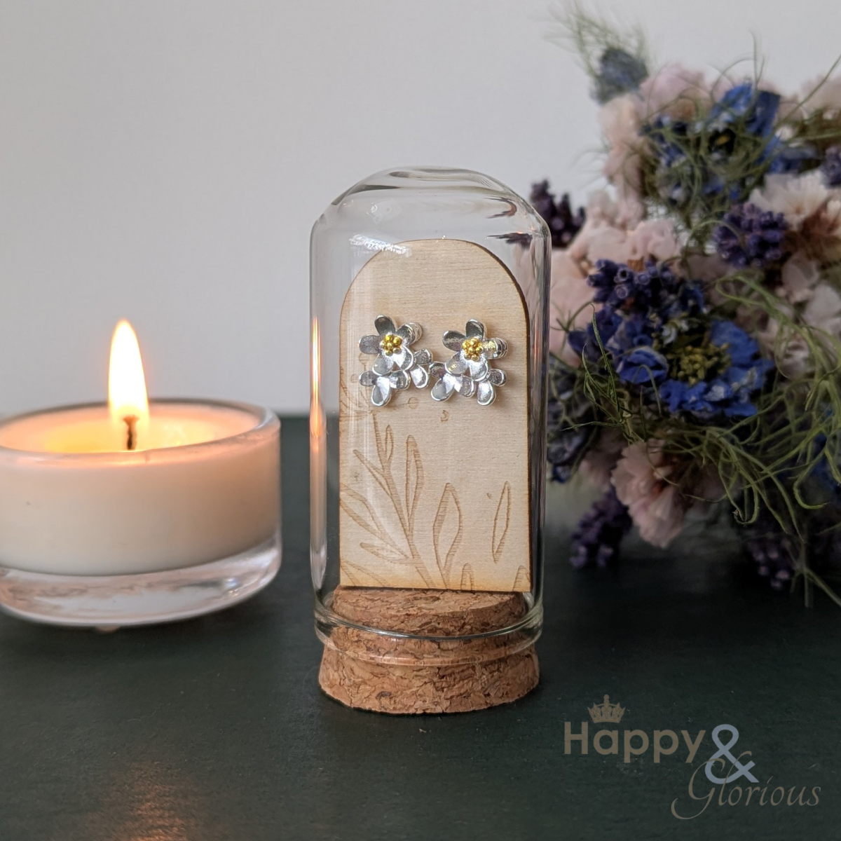 Silver earrings in a glass bottle with cork lid, next to a lit candle and flowers on a dark surface.