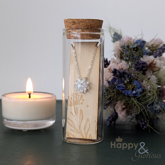 Necklace in a glass bottle with a wooden label, next to a lit candle and flowers on a dark surface.