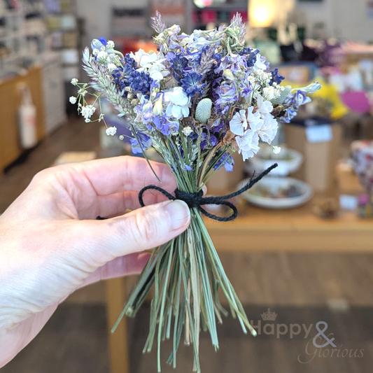 Hand holding a small bouquet of purple and white flowers with a blurred indoor background.